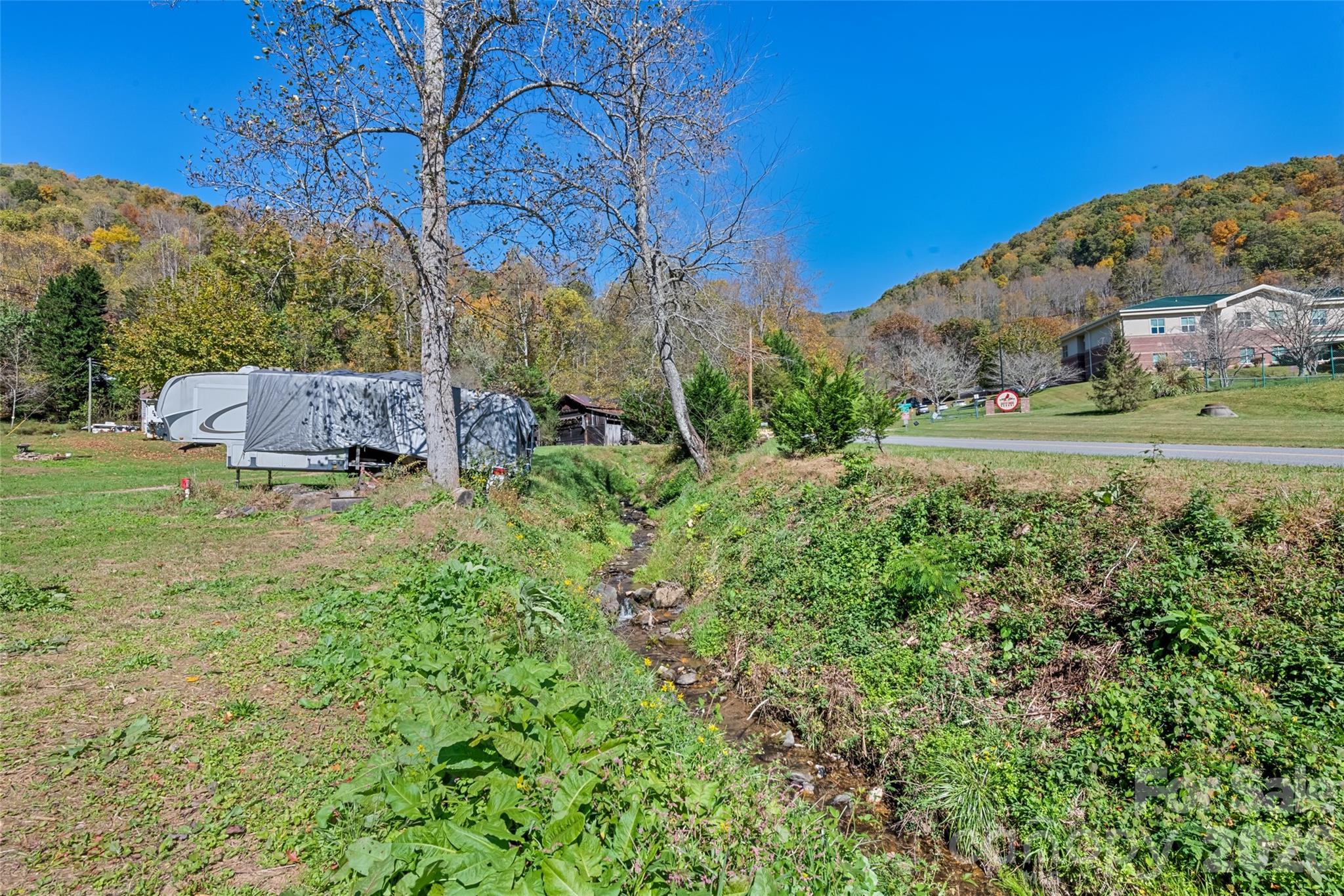 Grassy property with an RV beside a creek, backed by autumn-colored wooded hills.