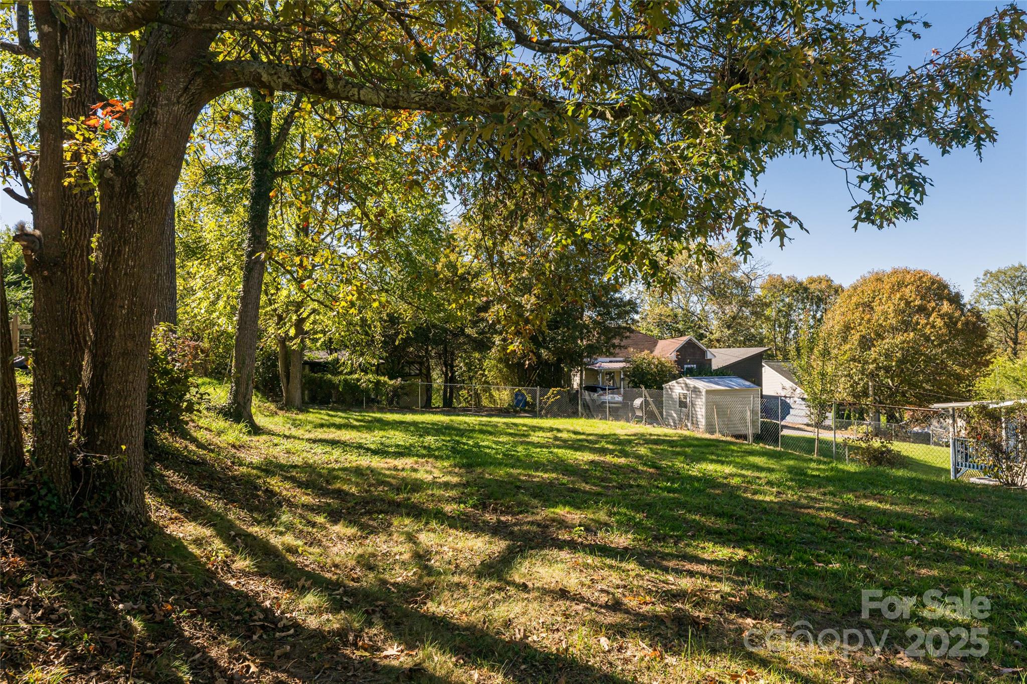Grassy residential lot shaded by mature trees, with a chain-link fence and neighboring homes beyond.