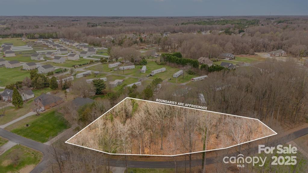 Aerial view of a wooded vacant lot outlined beside a road, with homes nearby.