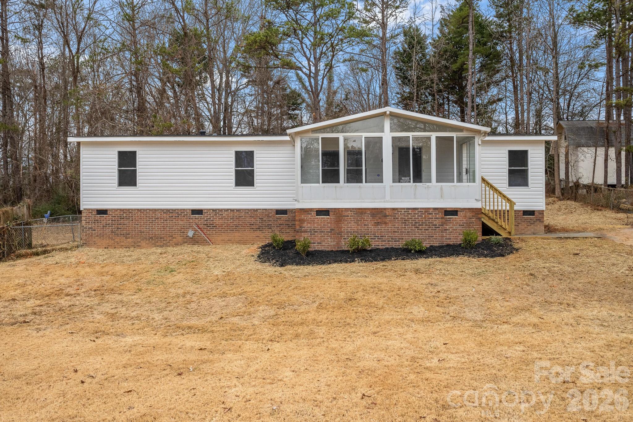White manufactured home with a brick foundation, enclosed sunroom, and wooden entry stairs.