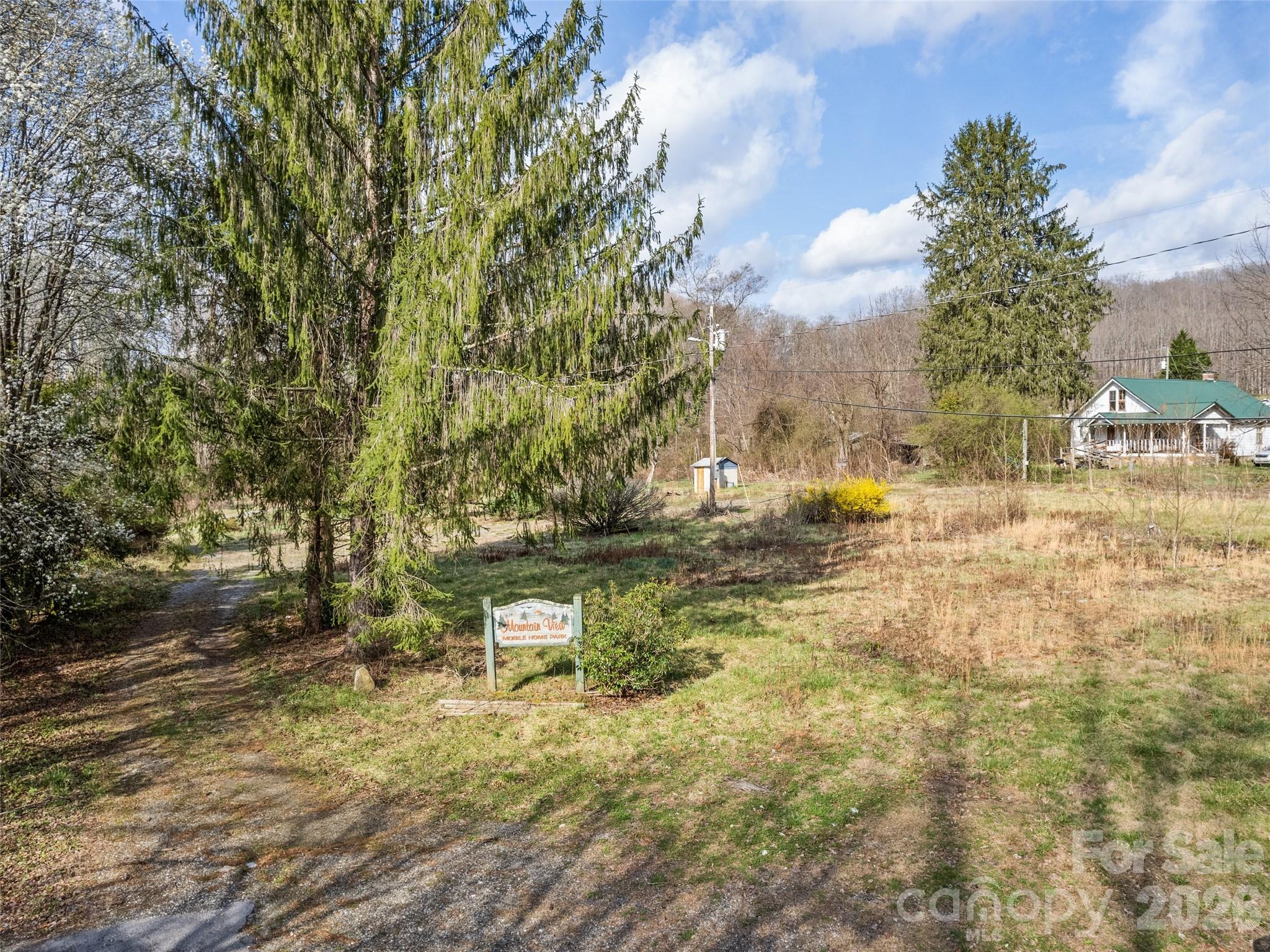 Mobile home park entrance with a Mountain View sign, gravel drive, large evergreen, and a distant white house.