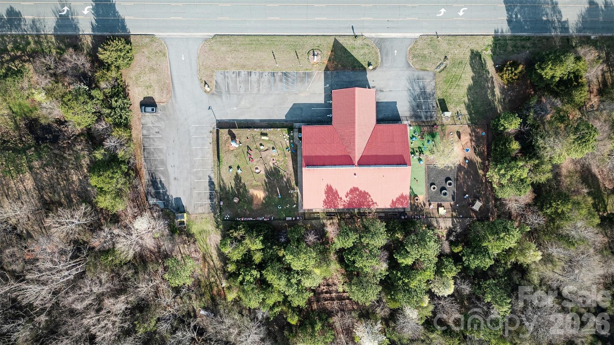 Aerial view of a red-roofed building with a parking lot and fenced play area among trees.