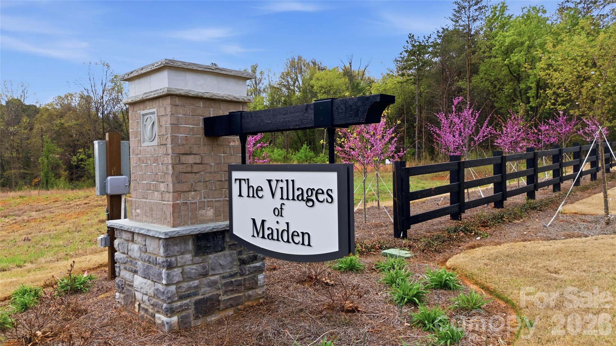 The Villages of Maiden entrance sign beside a black fence and blooming pink trees.