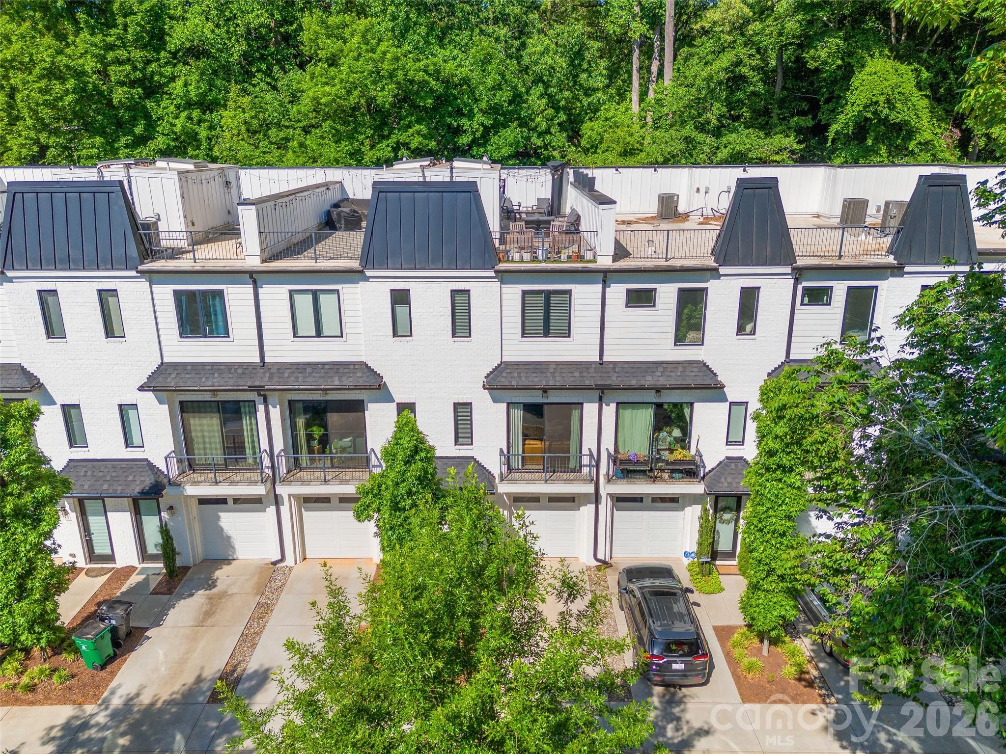 Row of white townhomes with dark roofs, balconies, garages, and mature trees.