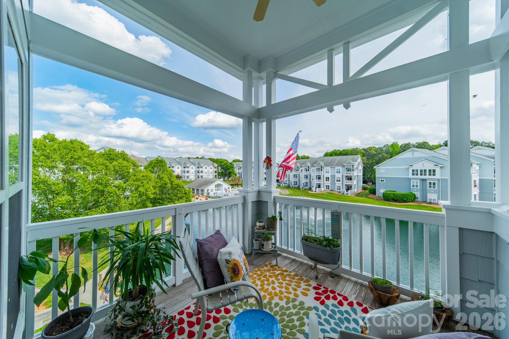 Apartment balcony with potted plants and a colorful rug overlooking a lake and nearby buildings.
