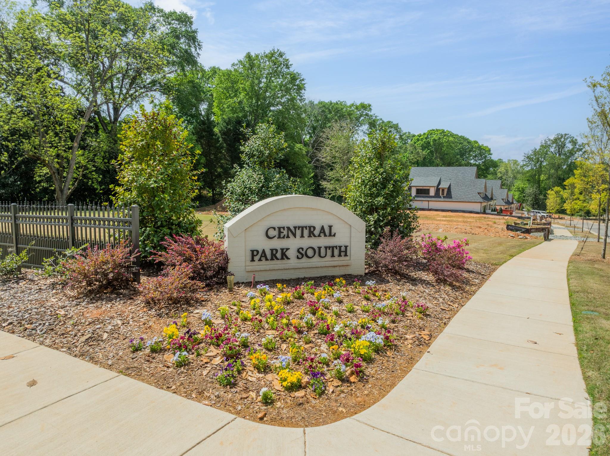 Central Park South entrance sign amid flower beds, with homes and a sidewalk in the background.