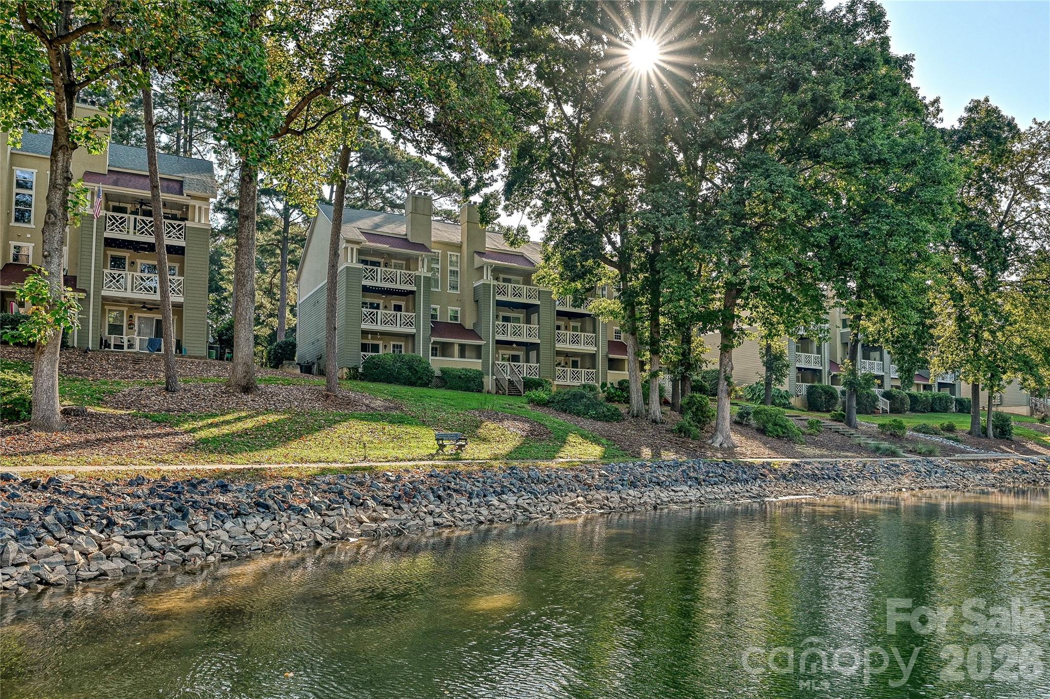 Residential building with balconies beside a tree-lined pond and rocky shoreline.