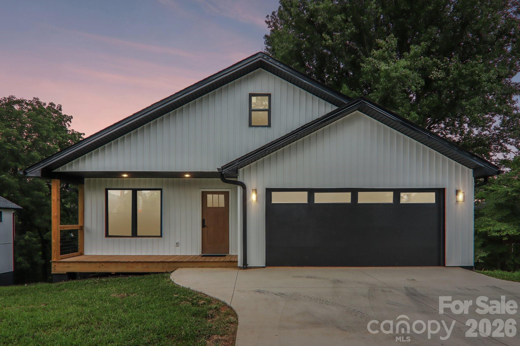 Modern gray house at dusk with a wooden porch, dark garage door, and concrete driveway.