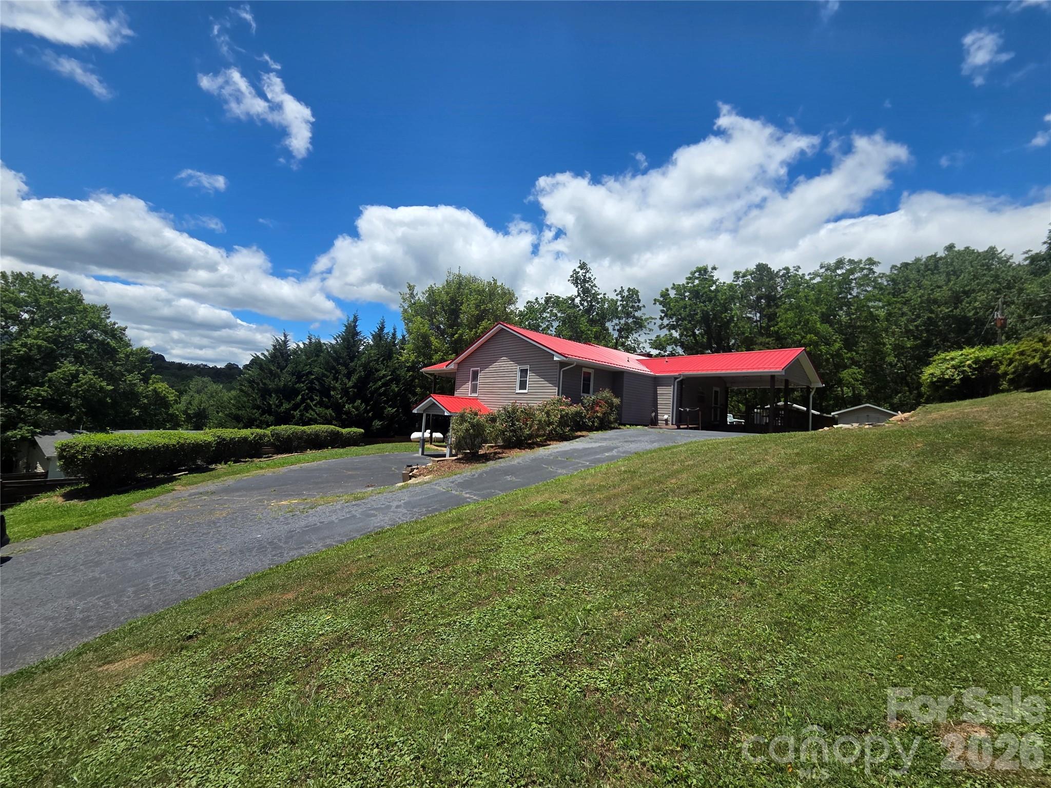 Gray-sided house with a red metal roof and covered carport beside a sloping lawn and paved driveway.