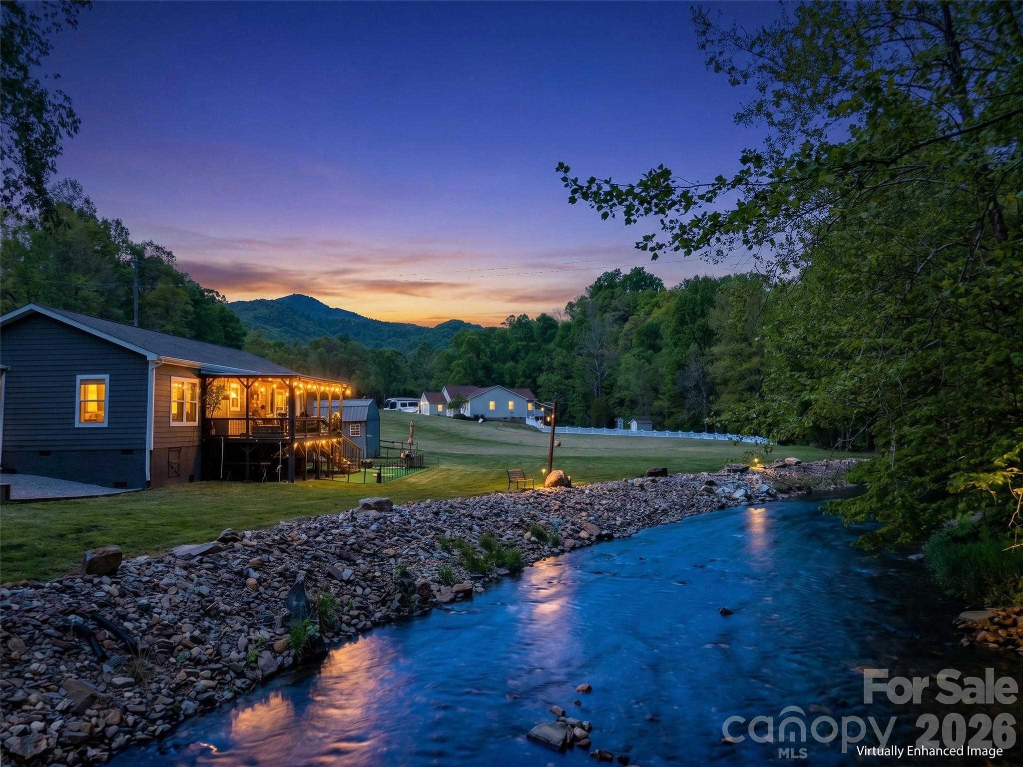 Riverside home with a warmly lit porch, rocky shoreline, and wooded hills at dusk.