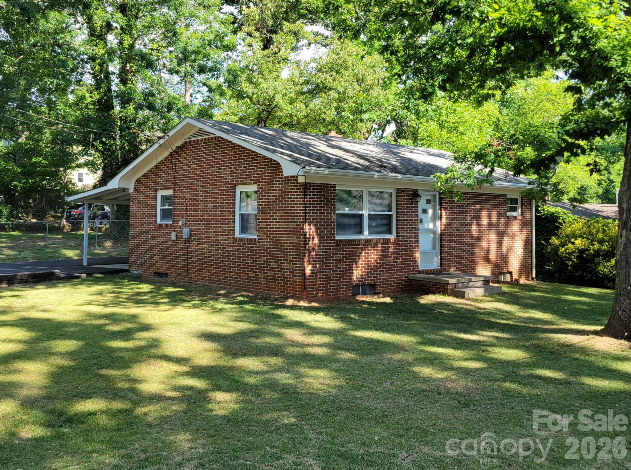 Single-story red brick house with white trim, a carport, and a shaded lawn.