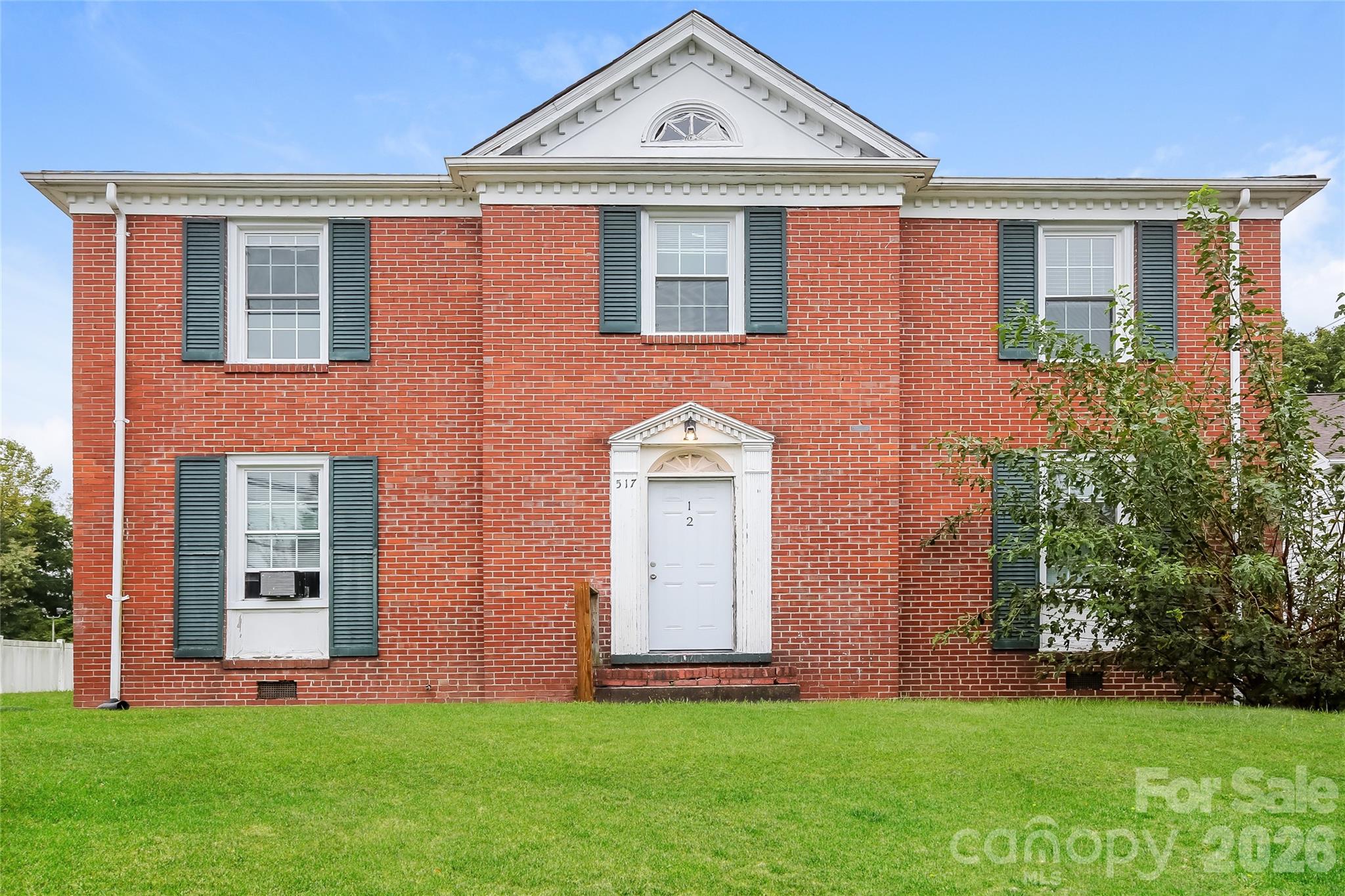 Two-story red brick home with white gable trim, dark shutters, and a leafy tree beside the lawn.