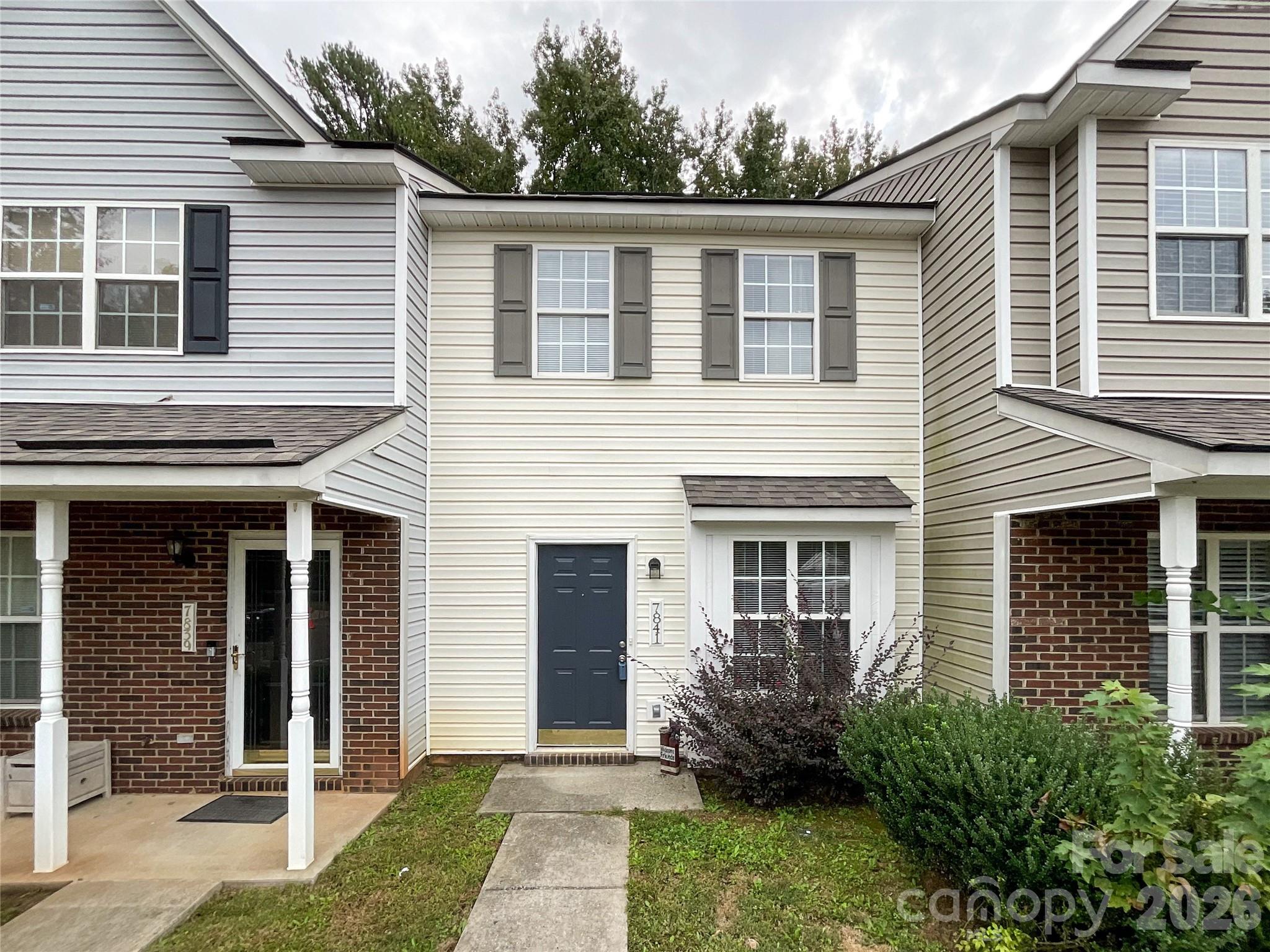 Townhouse front with cream siding, a dark blue door, brick porch sections, and shrubs.