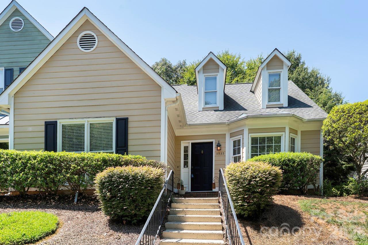 Beige-sided house with two roof dormers, a dark front door, front steps, and trimmed shrubs.