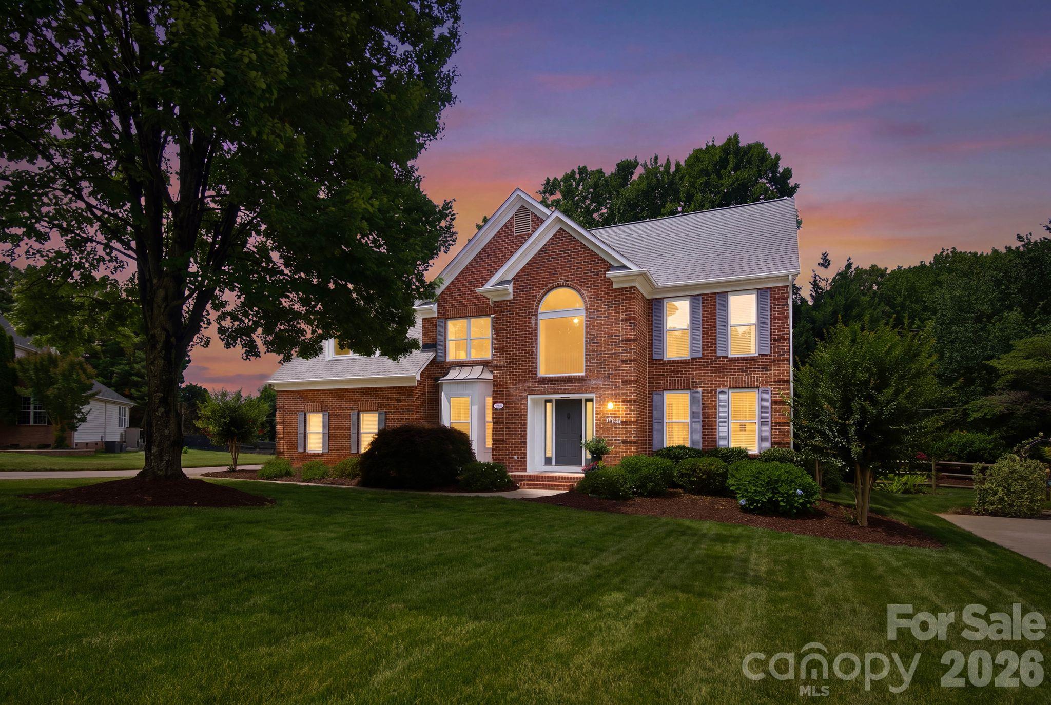 Two-story brick house with illuminated windows, landscaped shrubs, and a broad lawn at dusk.