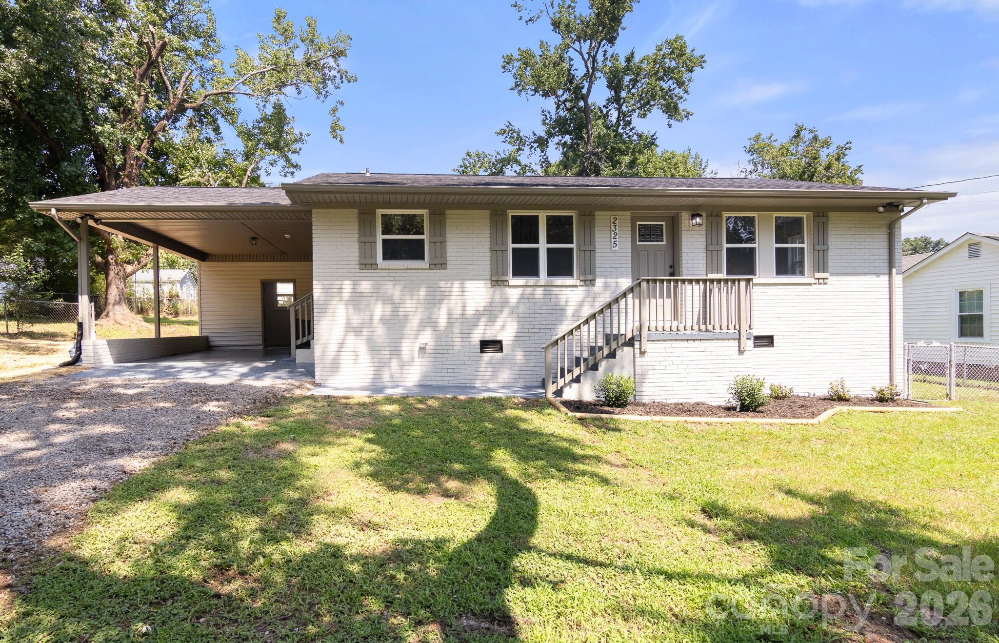 White brick house with a covered carport, raised front porch, and shaded grassy yard.