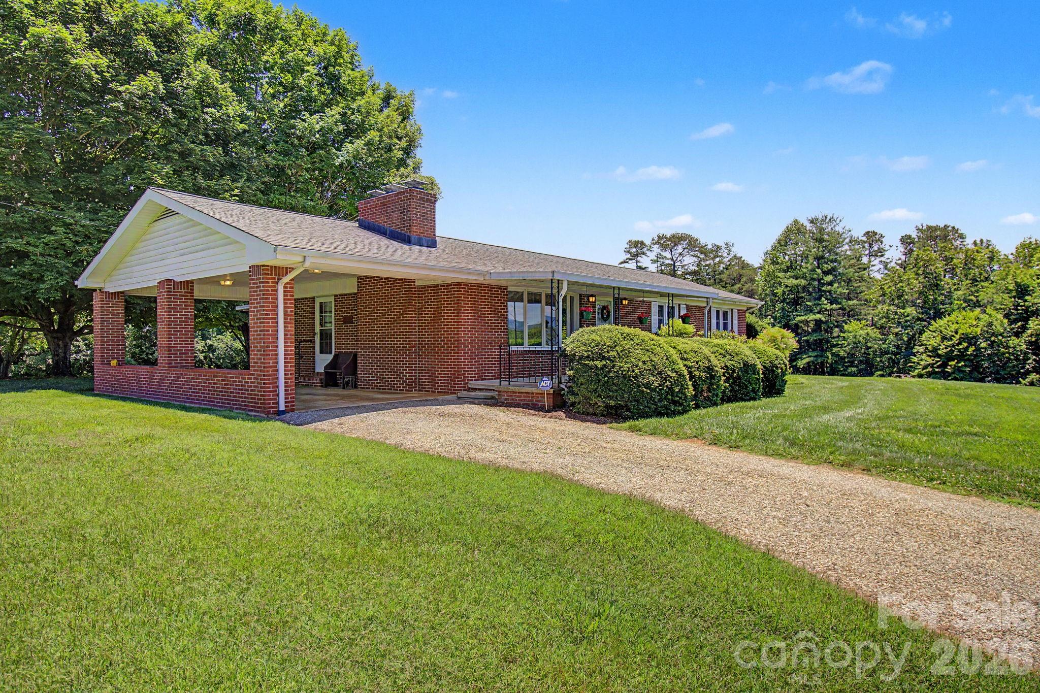 Single-story brick house with a white covered porch, chimney, and gravel driveway across a green lawn.