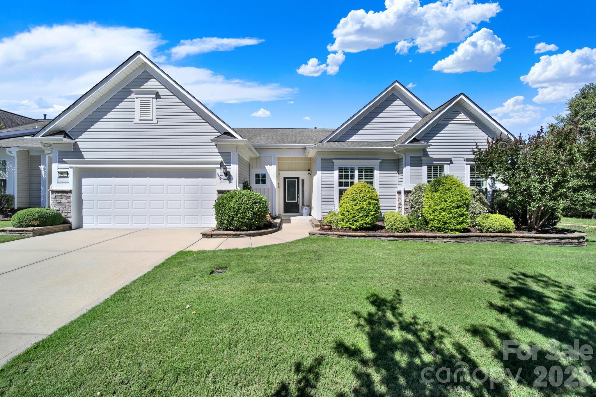 Gray single-story home with multiple gables, a two-car garage, landscaped shrubs, and a broad front lawn.