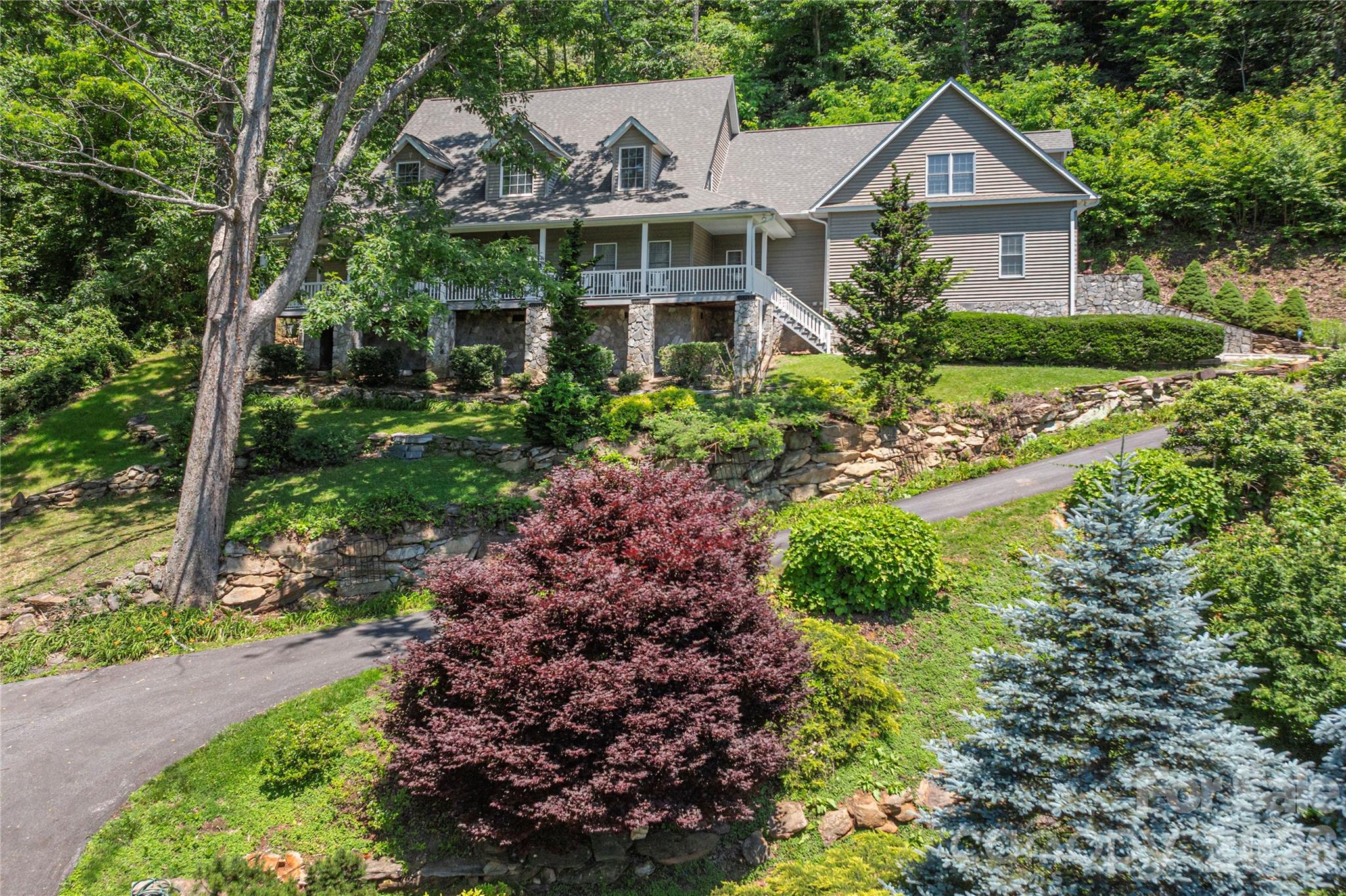 Gray multi-gabled house with a covered porch and stone foundation, surrounded by mature trees and landscaped grounds.