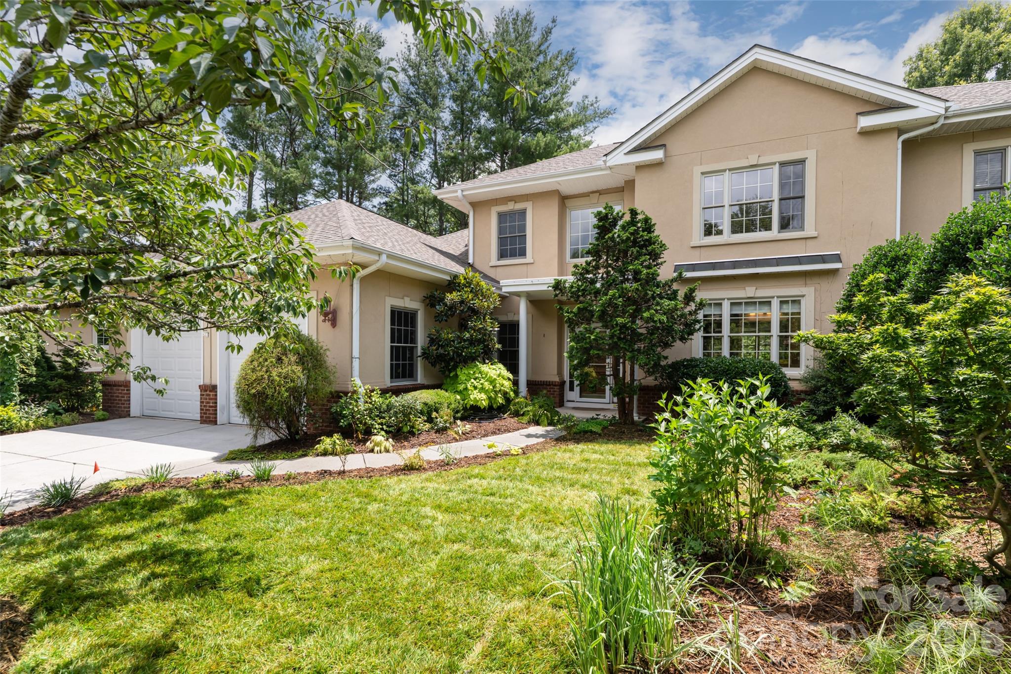 Two-story beige house with a two-car garage and a lush landscaped front yard.
