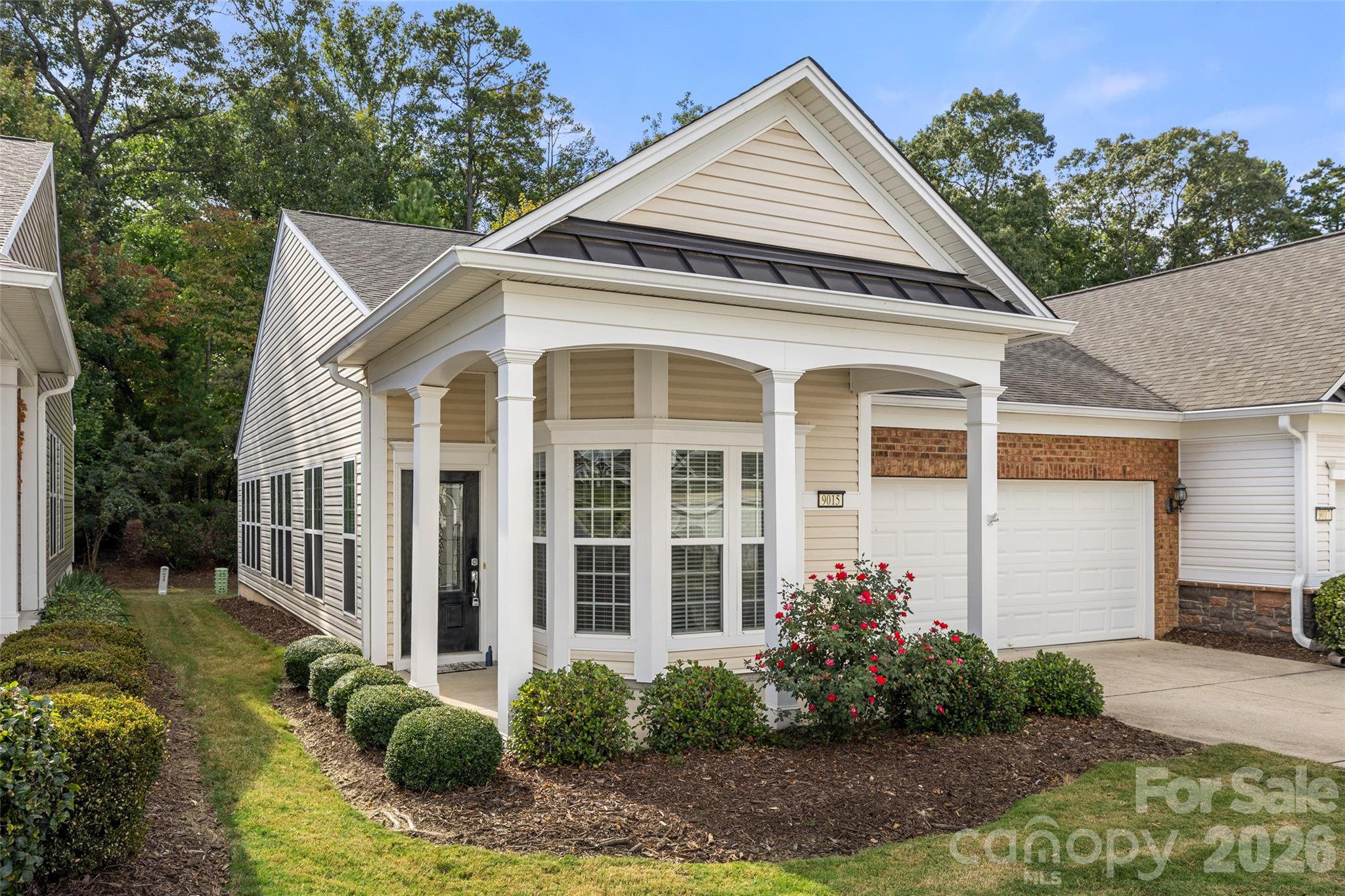 Beige home with a covered columned porch, bay windows, attached garage, and landscaped front yard.