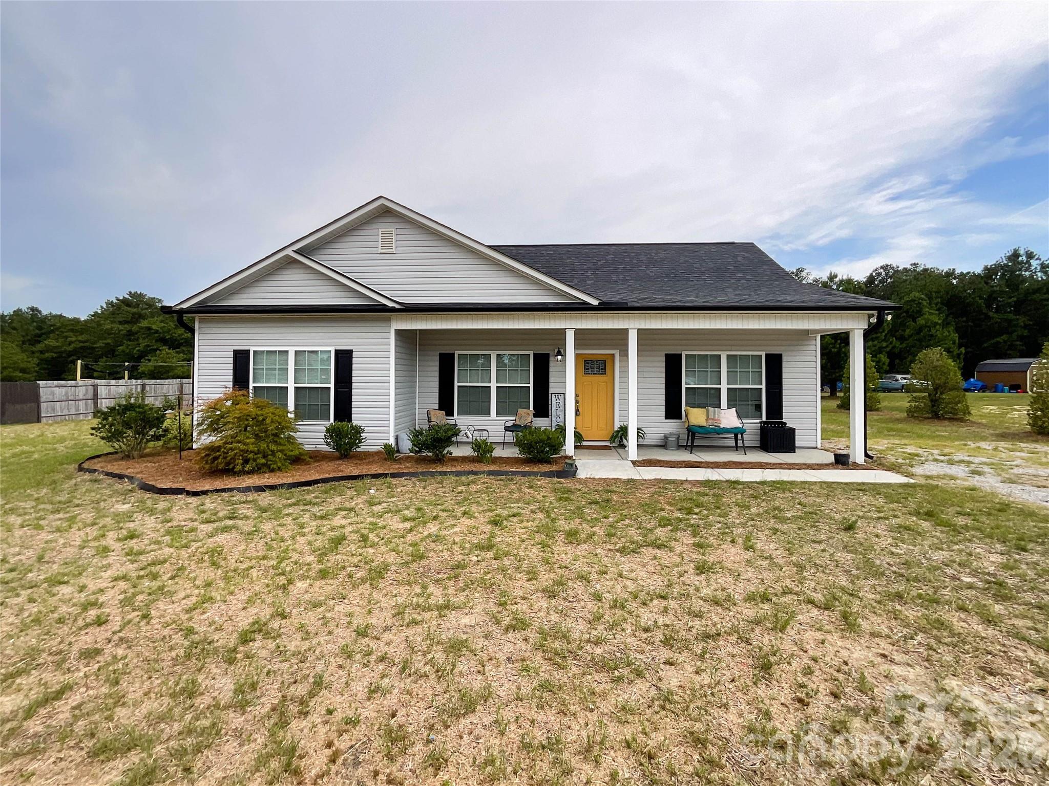 White single-story house with a covered porch, yellow front door, black shutters, and patchy lawn.