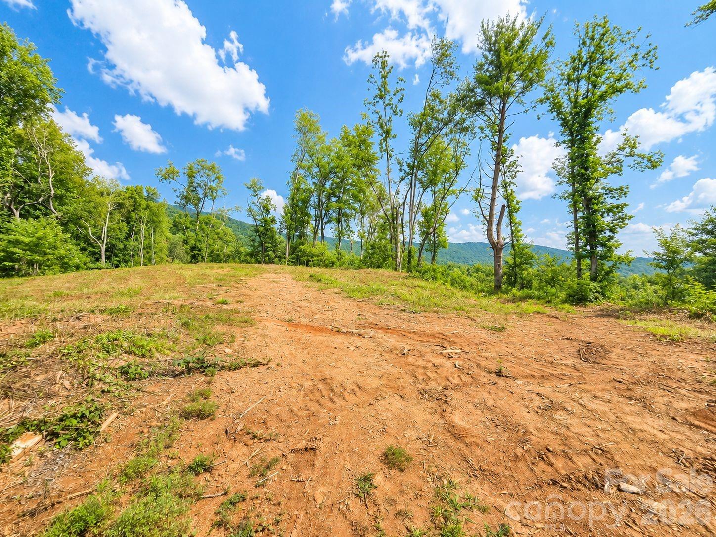 Cleared dirt lot edged by trees, with a wooded mountain ridge in the distance.