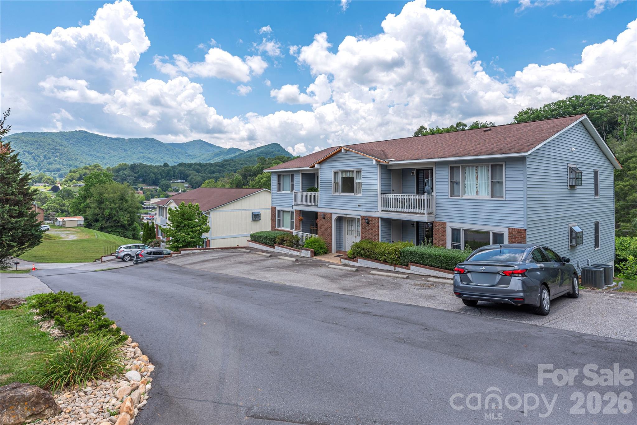 Blue-sided residential building with balconies and parking, set against wooded mountain ridges.