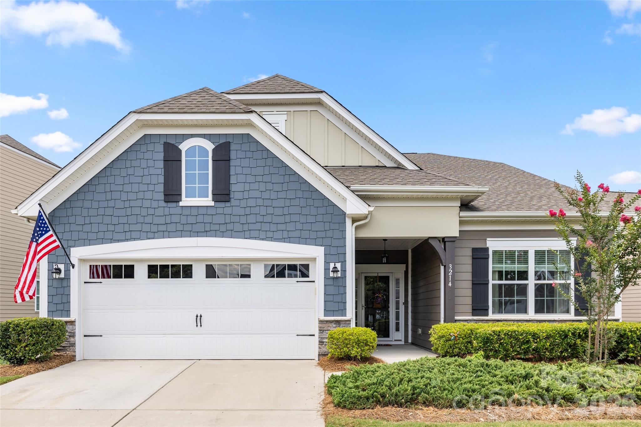 Two-story blue-and-gray home with a white double garage, American flag, and landscaped front yard.