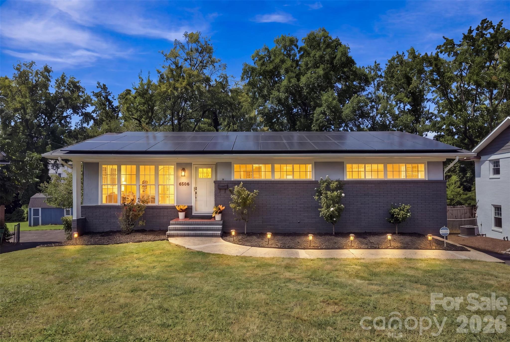 Single-story home with rooftop solar panels, dark brick facade, and warmly glowing windows at dusk.