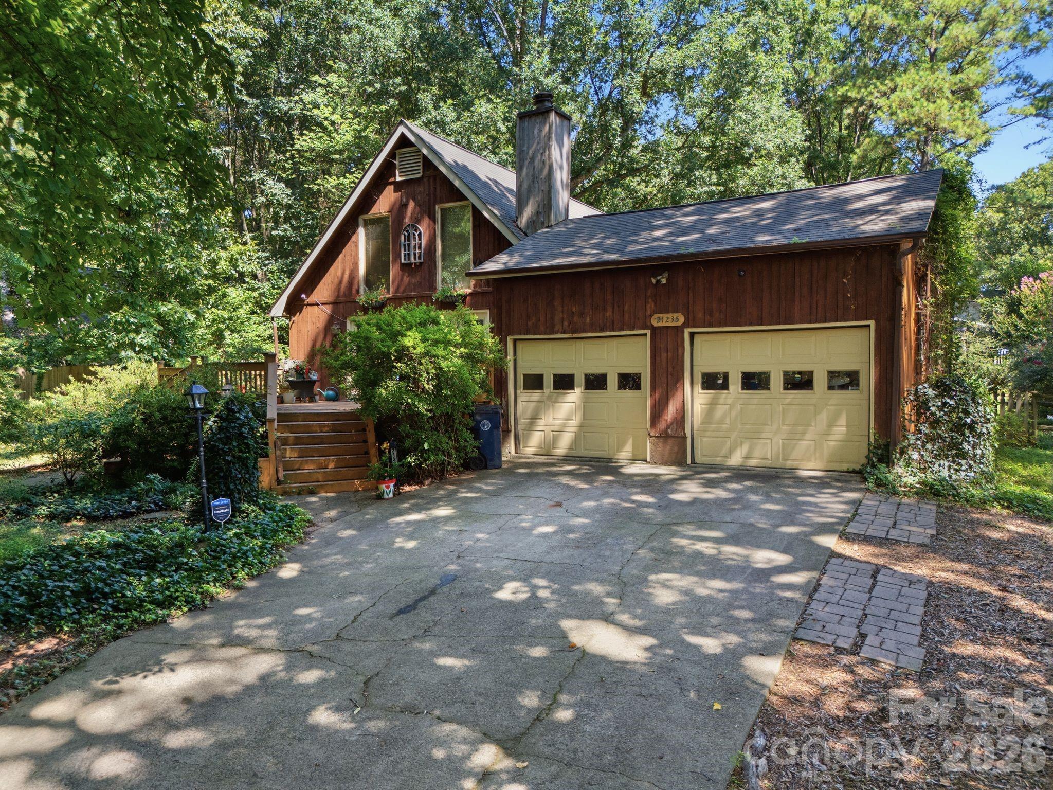 Wood-sided house with two pale yellow garage doors, a broad driveway, and mature trees.