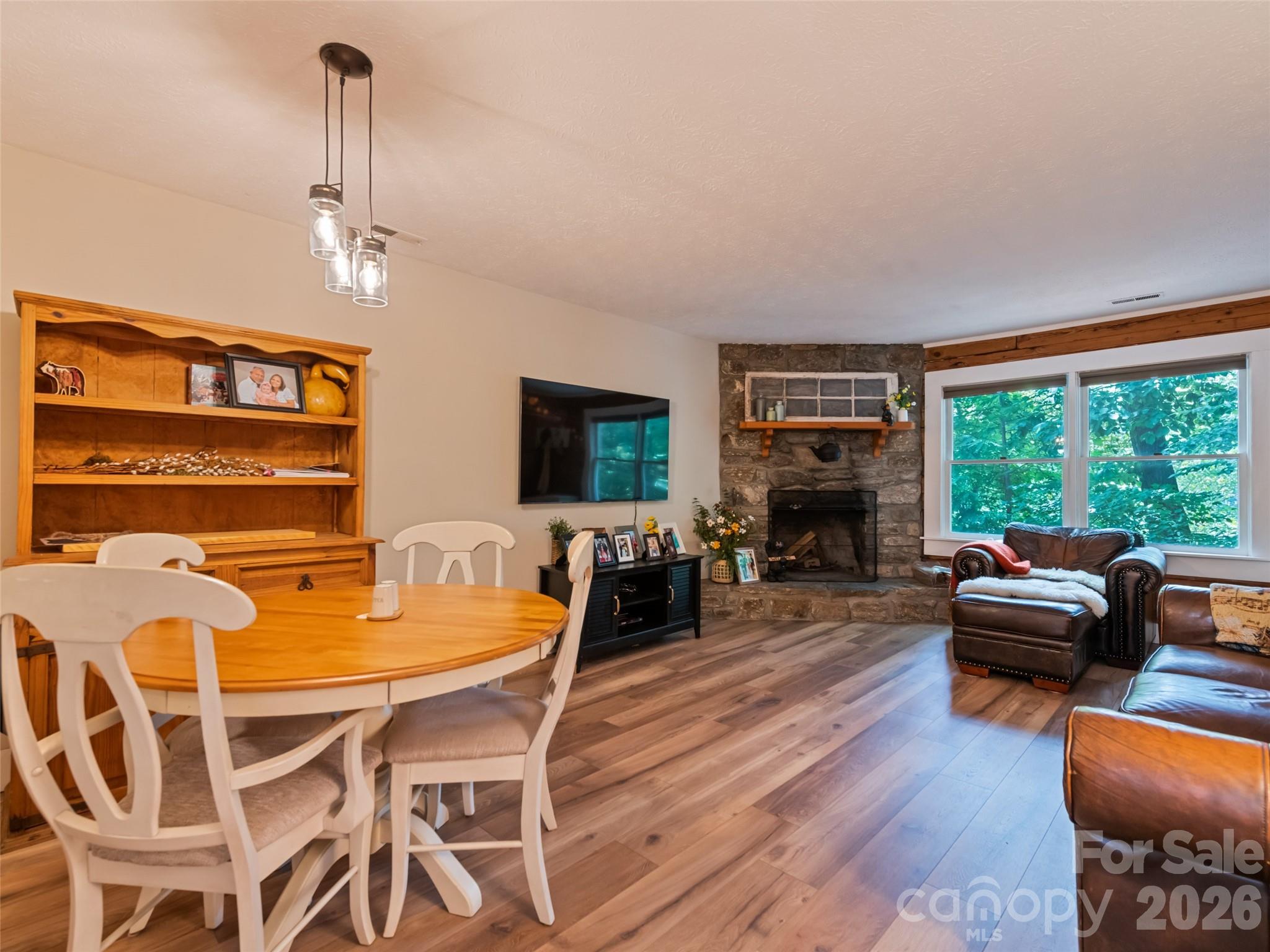 Living and dining room with a stone fireplace, leather seating, and large windows overlooking trees.
