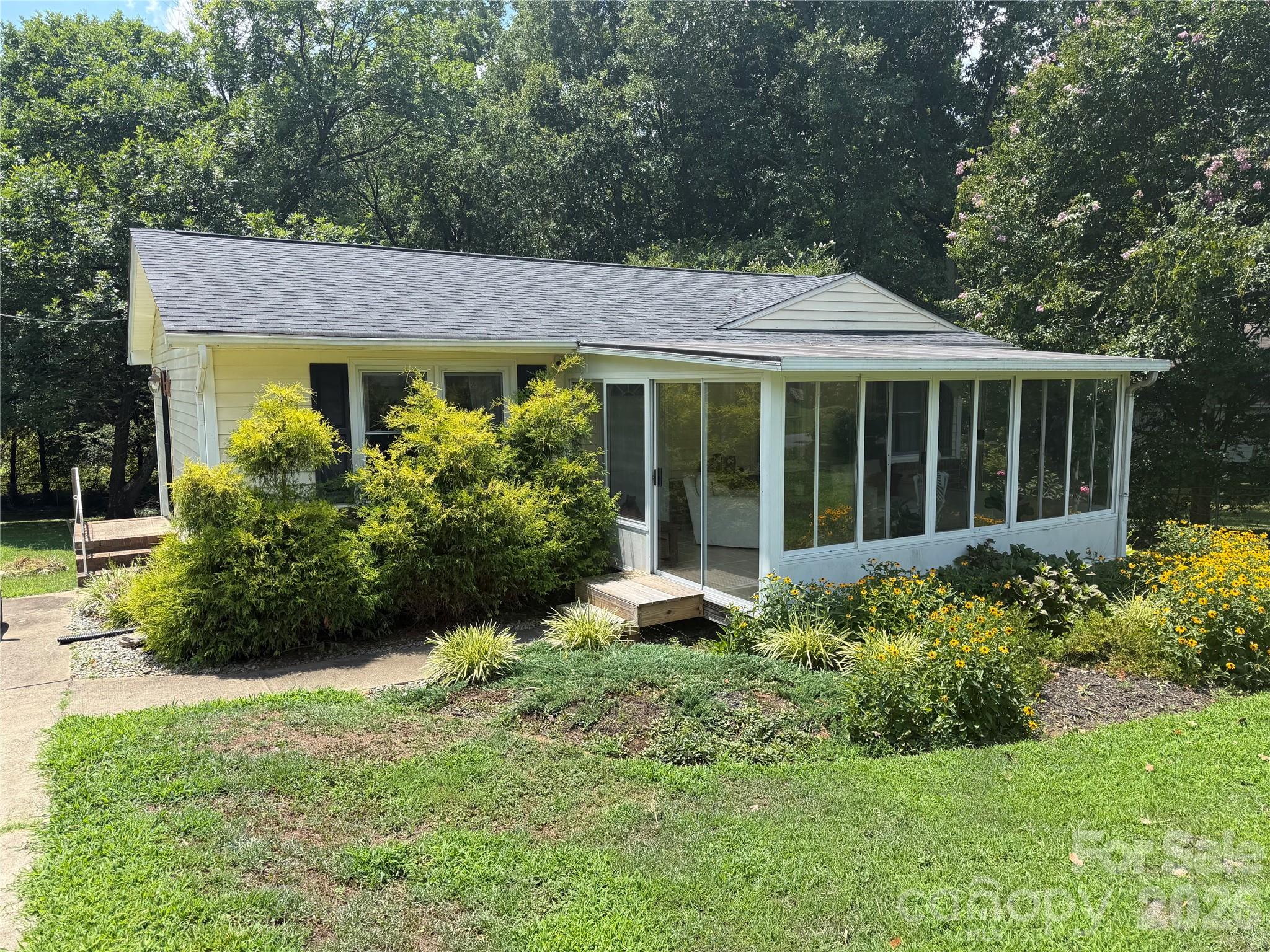 Single-story house with a glass-enclosed sunroom, flowering garden, and mature trees.