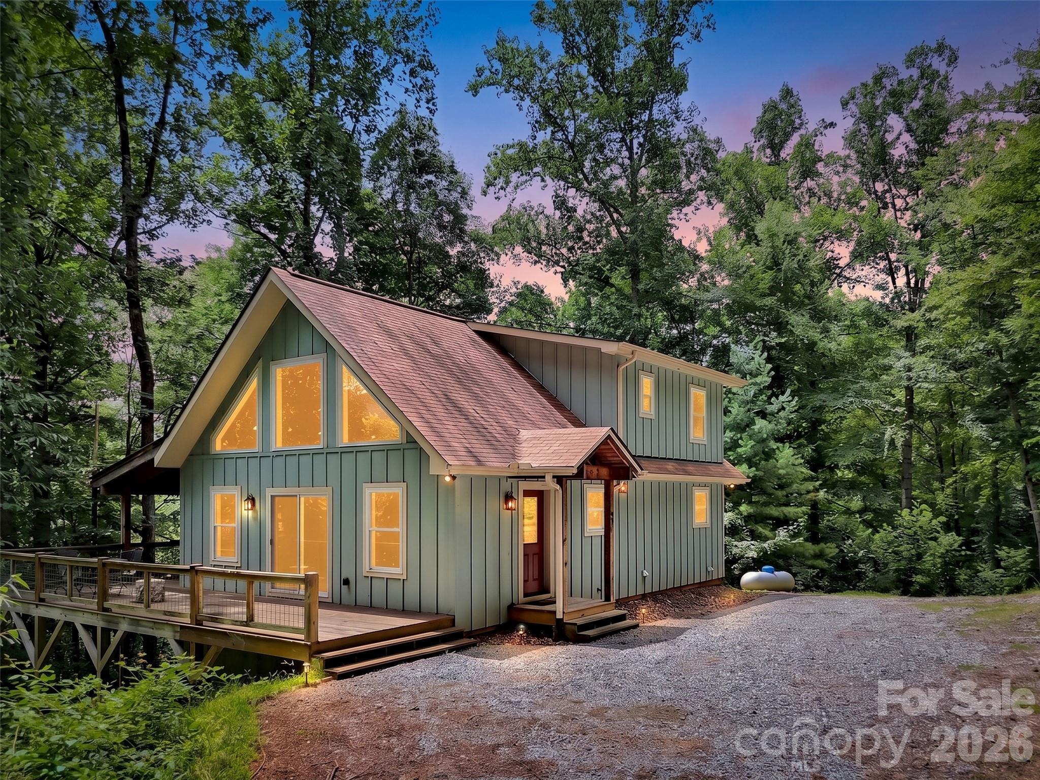 Green A-frame-style home with a raised deck, glowing windows, and a gravel driveway in the woods.