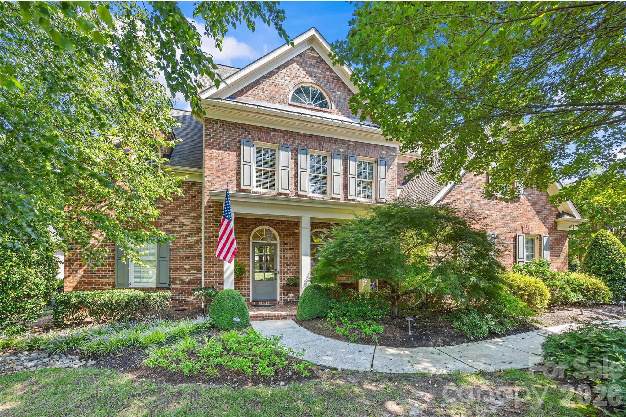 Two-story brick house with a covered entrance, American flag, and mature leafy landscaping.