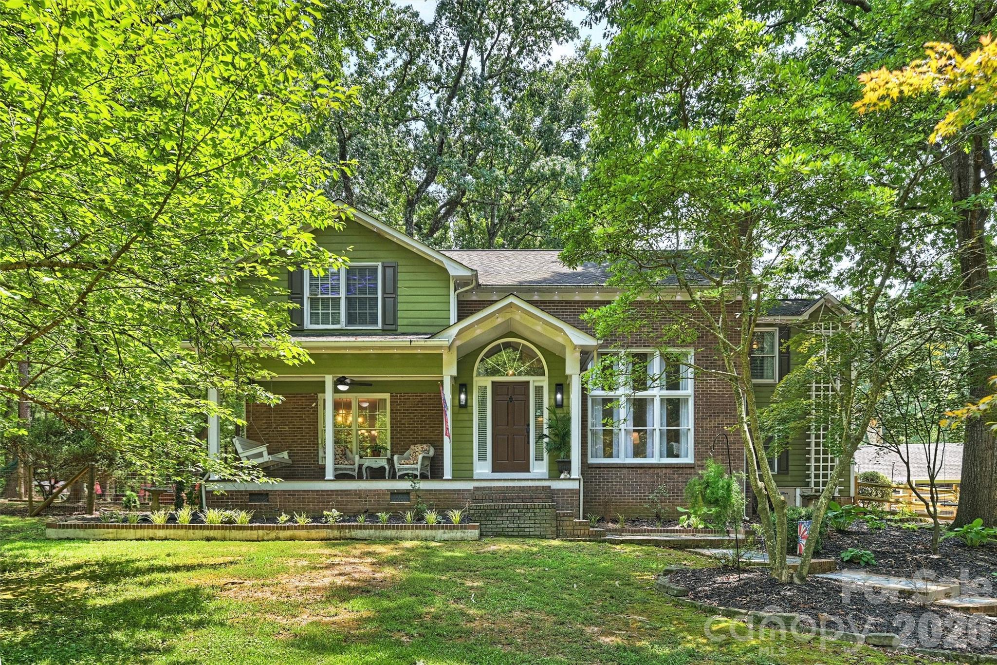 Two-story green-and-brick home with a covered porch, framed by mature trees.