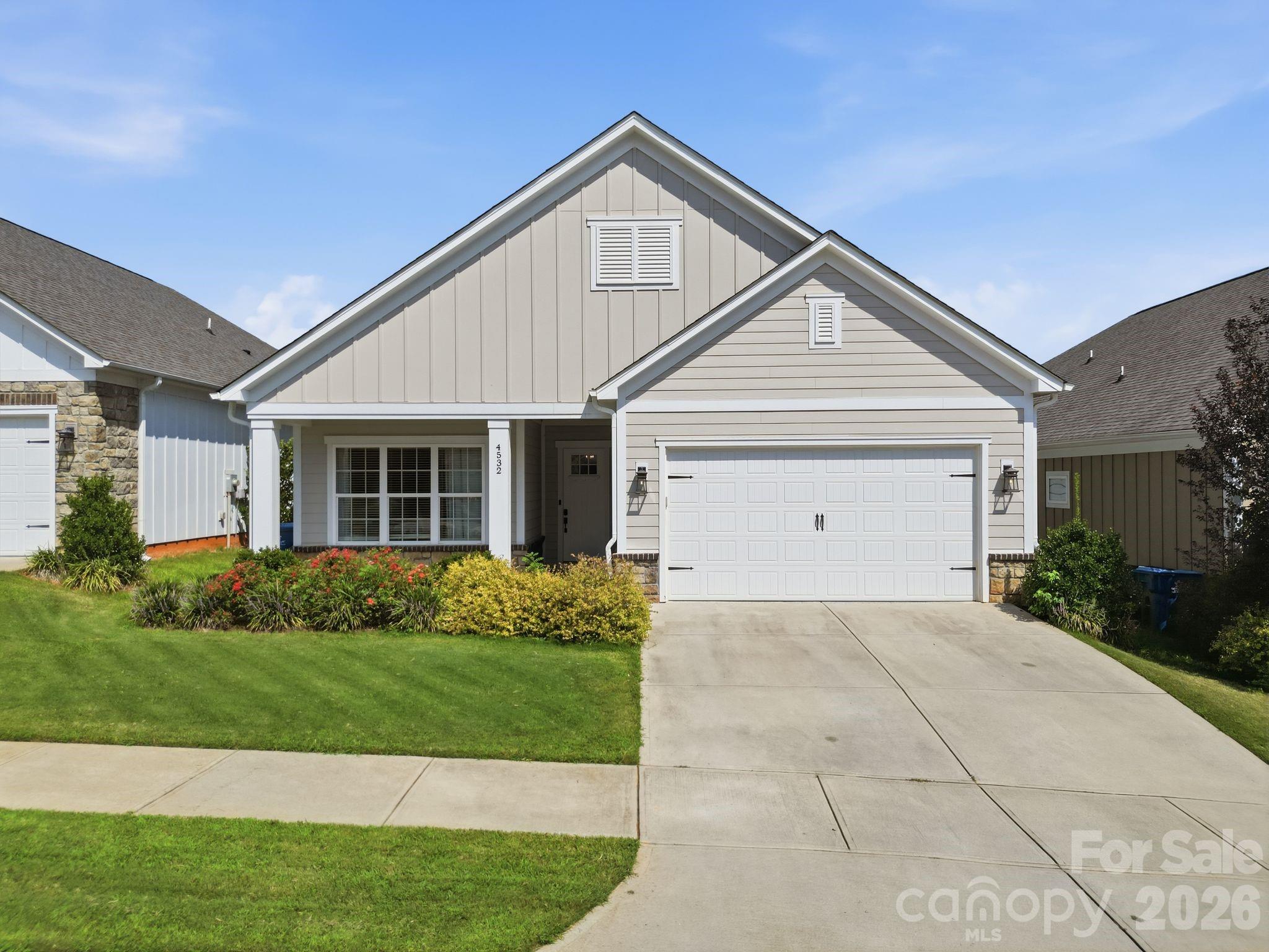Light-colored gable-front house with a two-car garage, landscaped flower beds, and a wide concrete driveway.