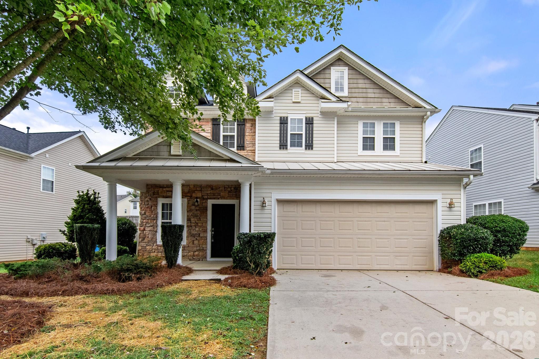 Two-story beige house with stone accents, a covered porch, and a two-car garage beneath a leafy tree.