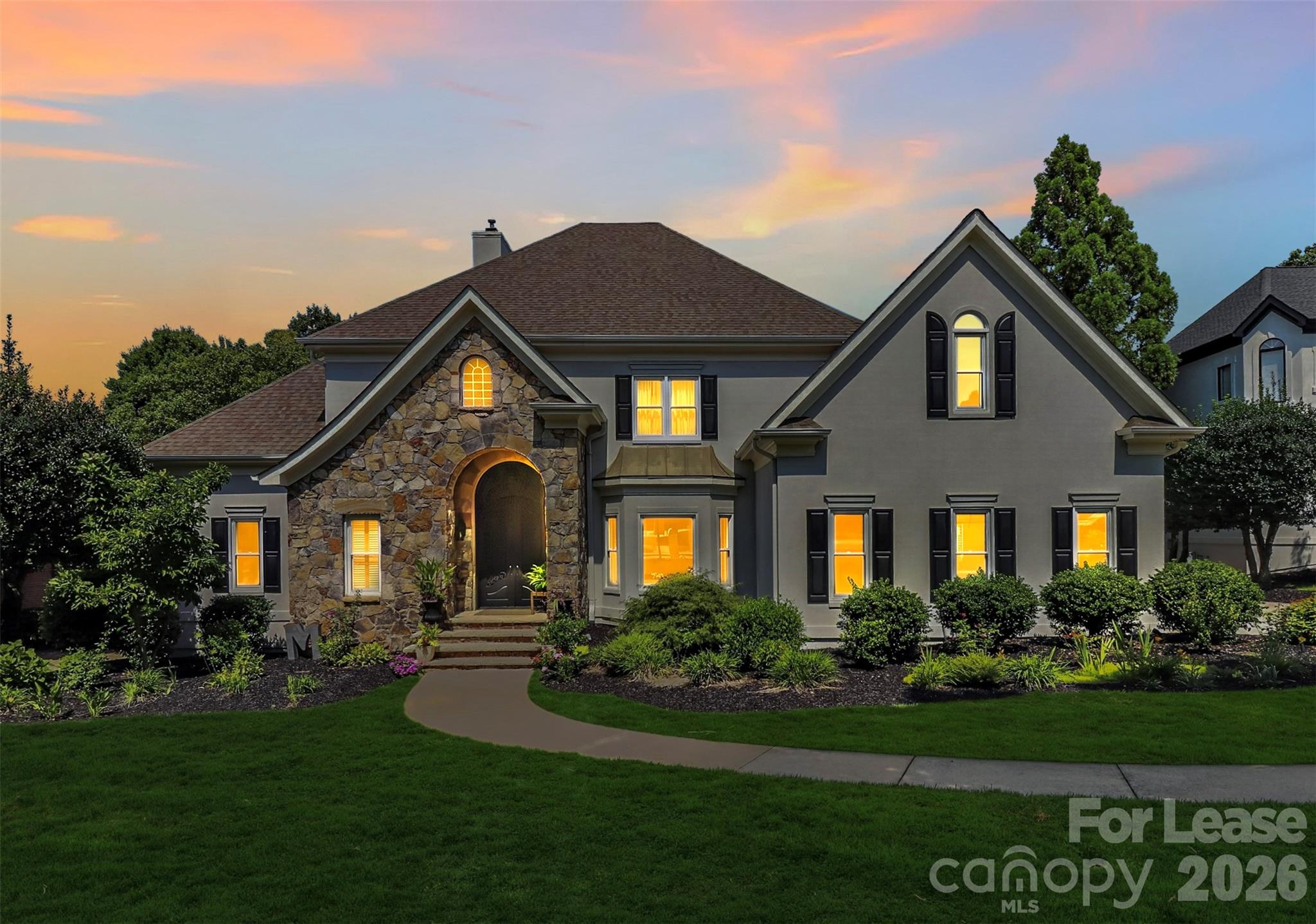 Gray two-story home with a stone entry, glowing windows, and landscaped front yard at dusk.