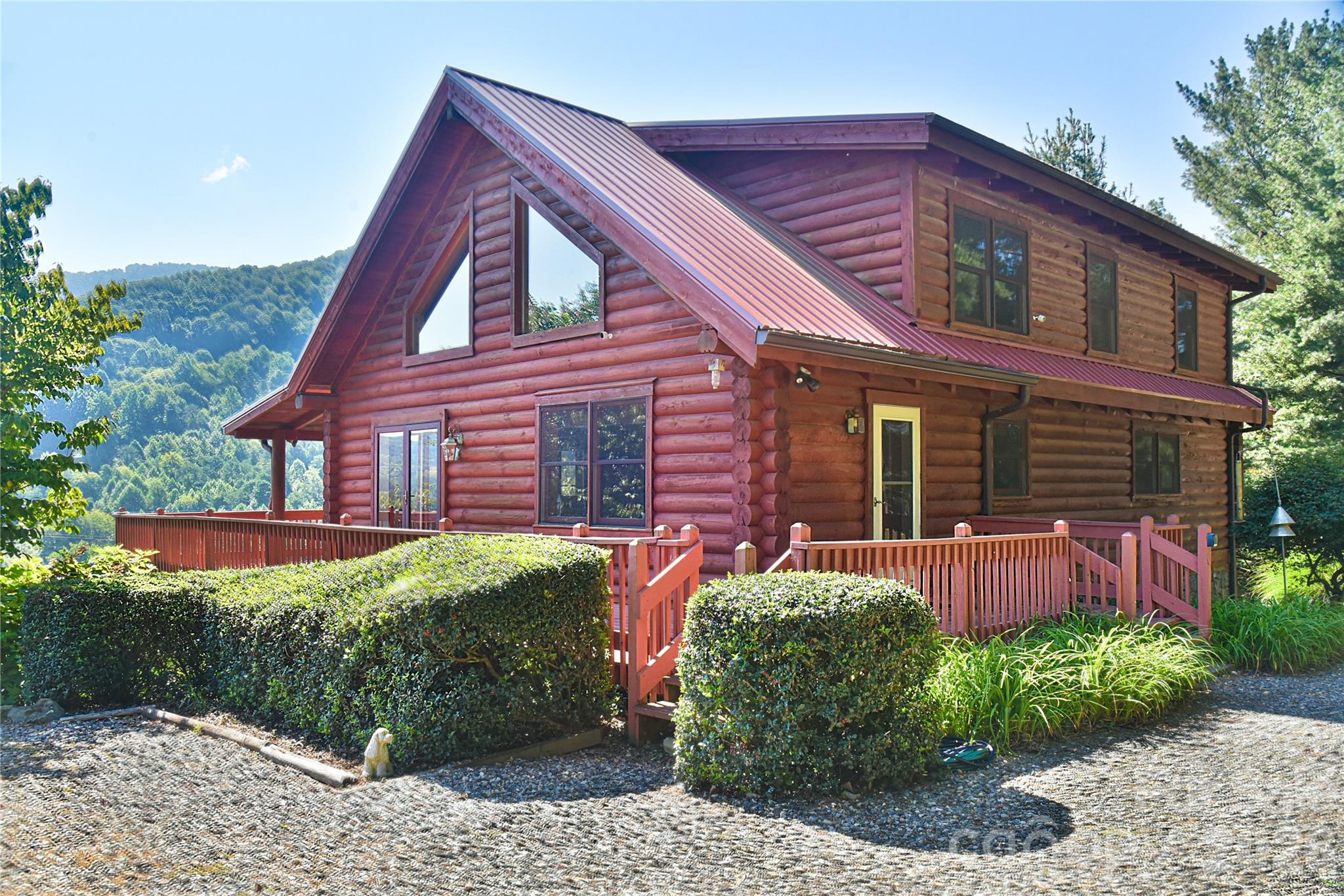 Two-story log cabin with a red deck, trimmed hedges, and wooded mountain views.
