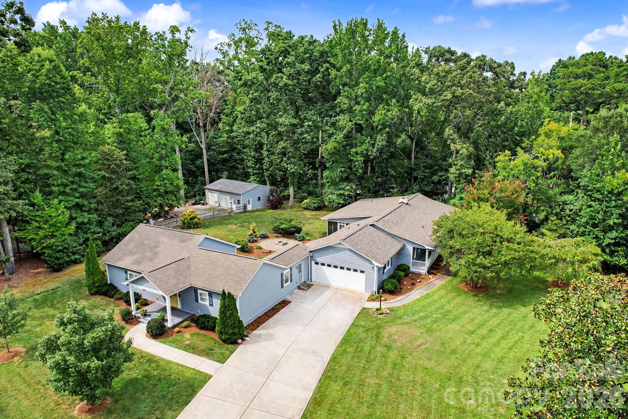 Aerial view of a blue-gray home with a broad driveway, landscaped lawn, and wooded surroundings.