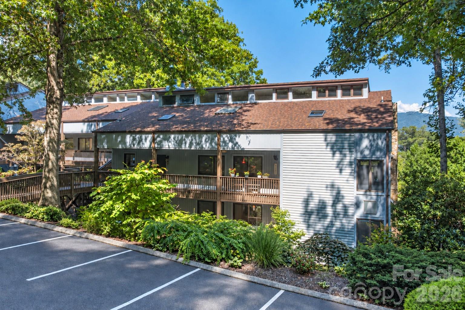 Multi-unit building with a long brown roof, wooden balcony, landscaped grounds, and parking spaces.