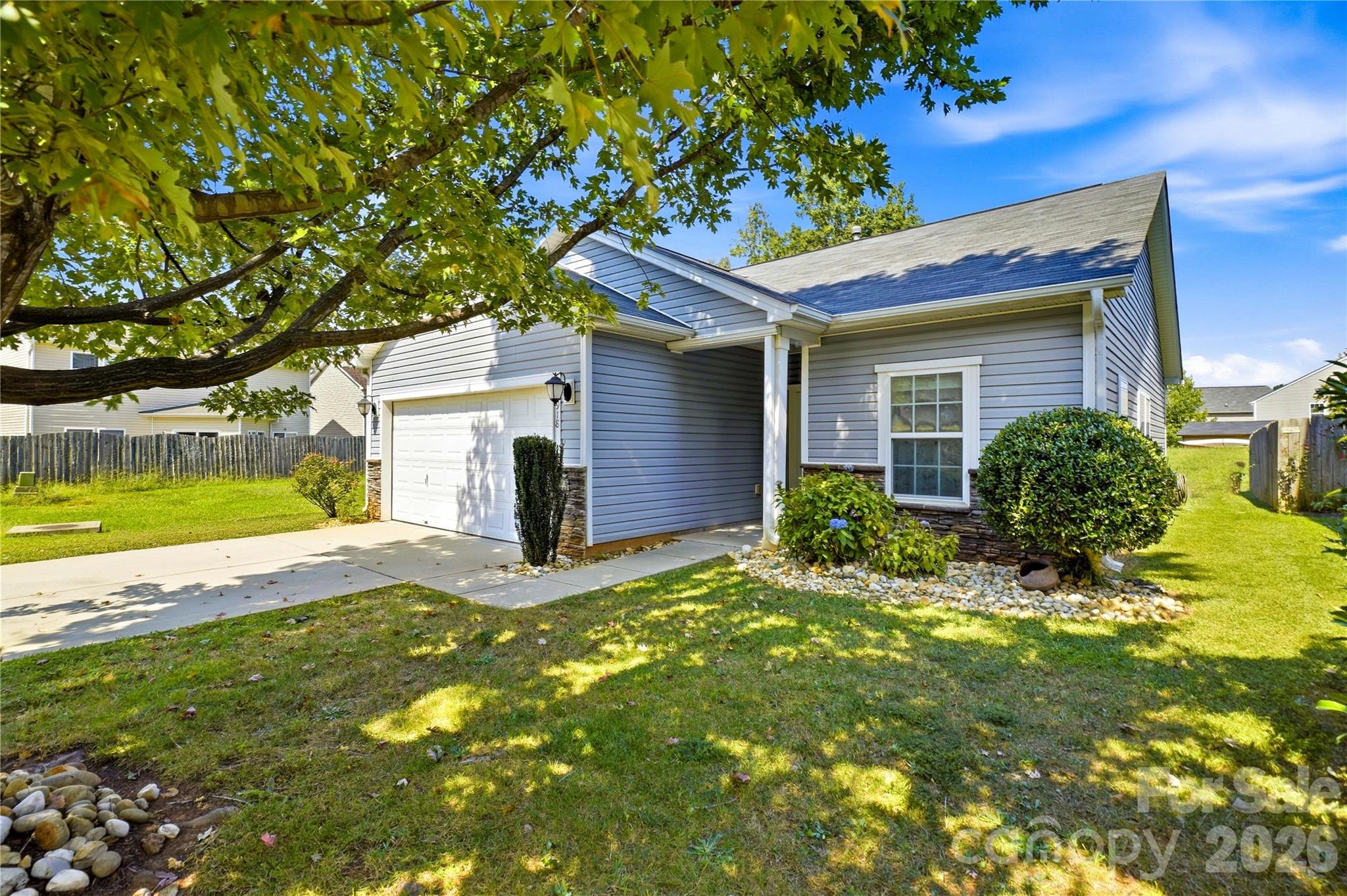 Gray-sided house with a white garage, front porch, and large shade tree over the lawn.