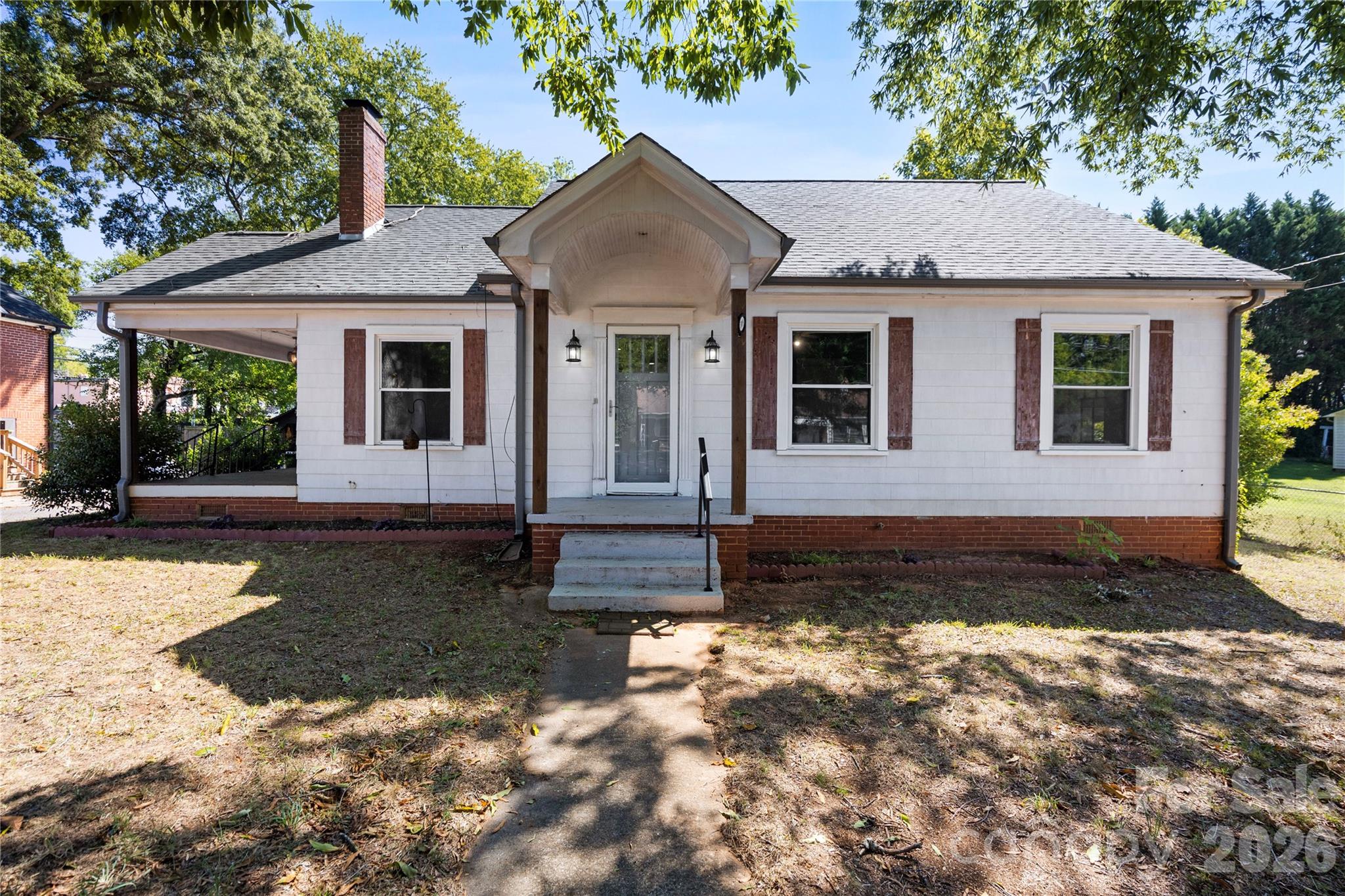 White single-story house with brick shutters and a covered entrance beneath mature trees.