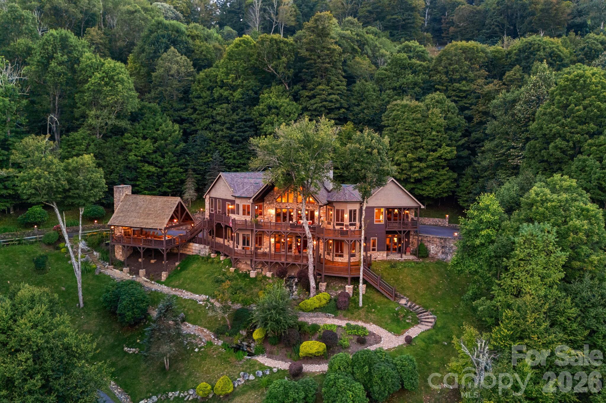 Wood-and-stone hillside home with wraparound decks, landscaped paths, and warm lights glowing at dusk.