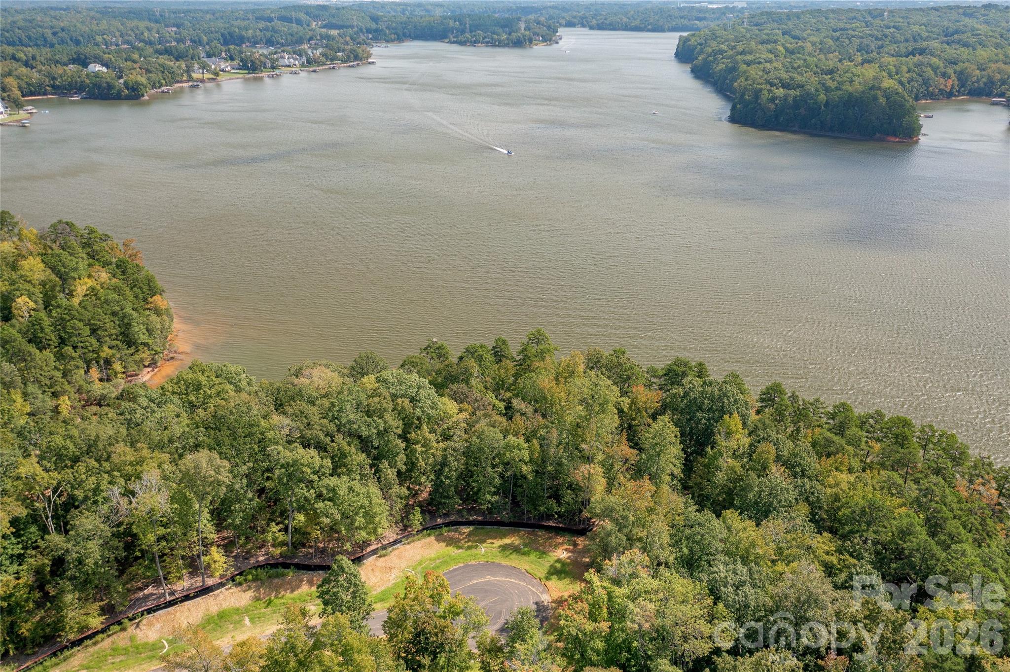 Aerial lake view with wooded shorelines, a boat wake, and a curved road among the trees.