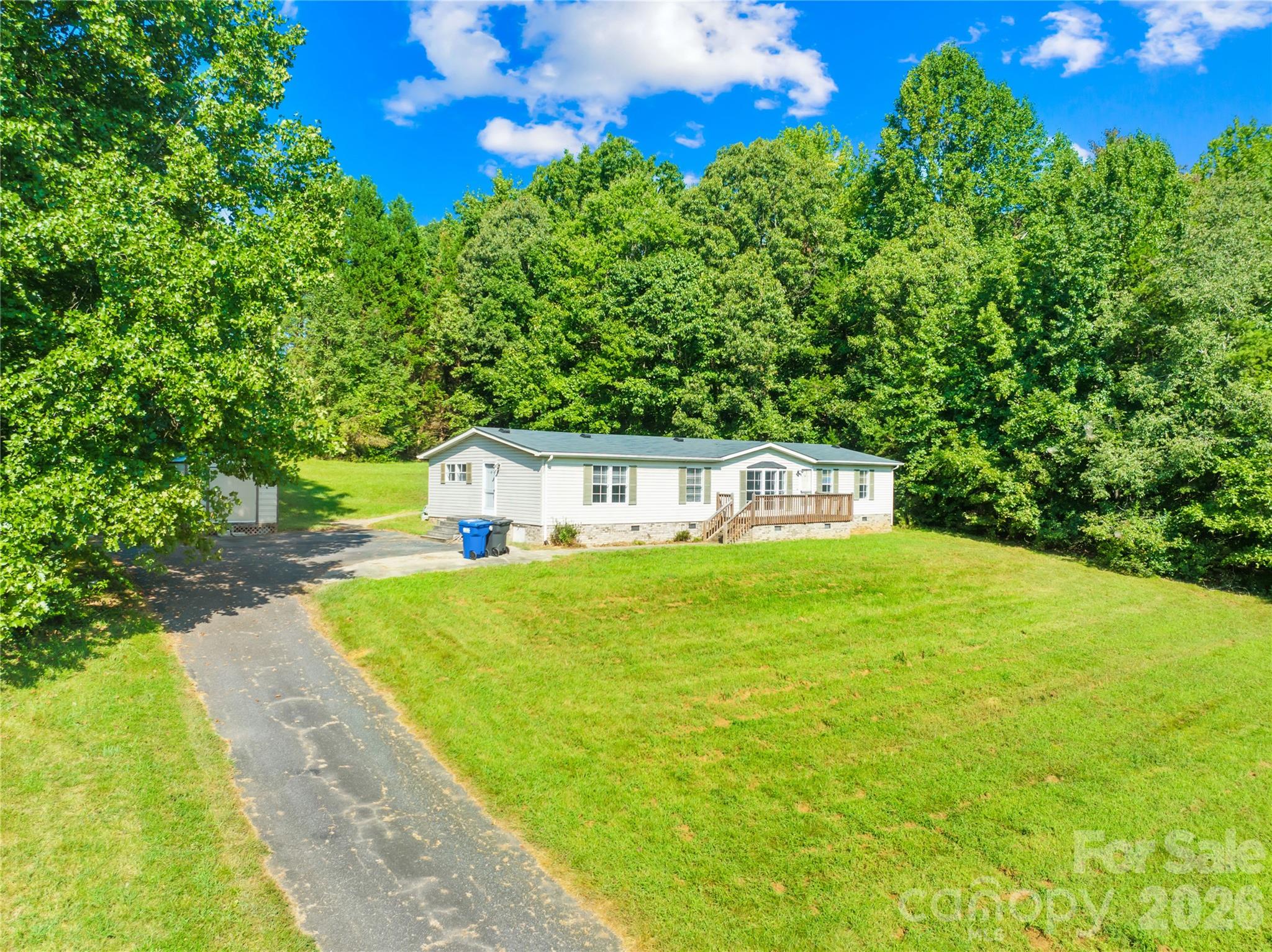 Single-story home with a wooden deck, broad lawn, curving driveway, and dense trees behind it.