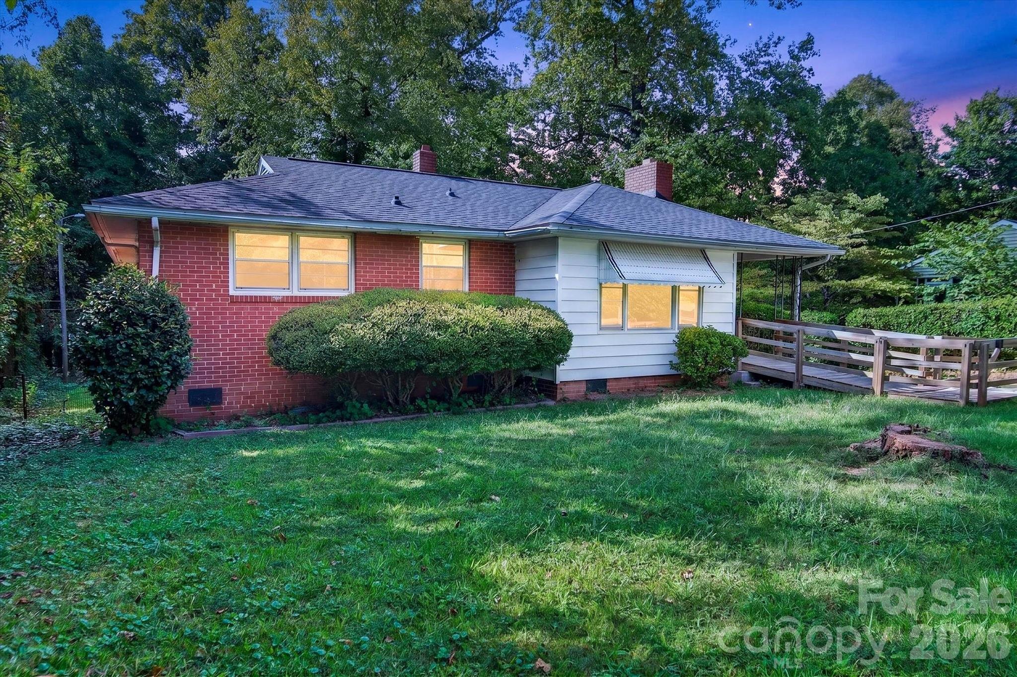 Single-story brick and white-sided house with lit windows, a wooden ramp, and a tree-lined lawn.