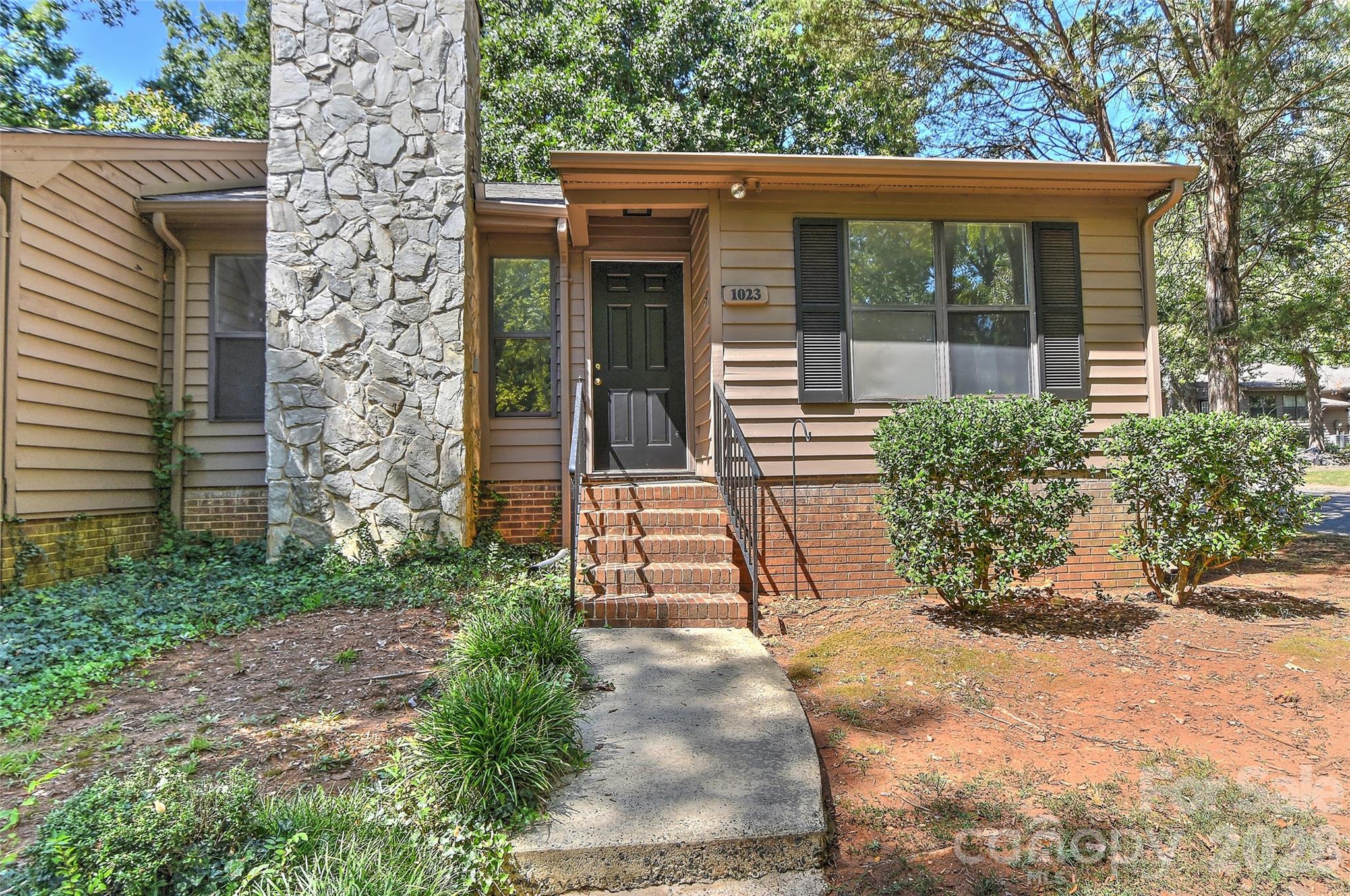 Tan-sided home with a prominent stone chimney and brick entry steps; house number 1023 beside the front door.