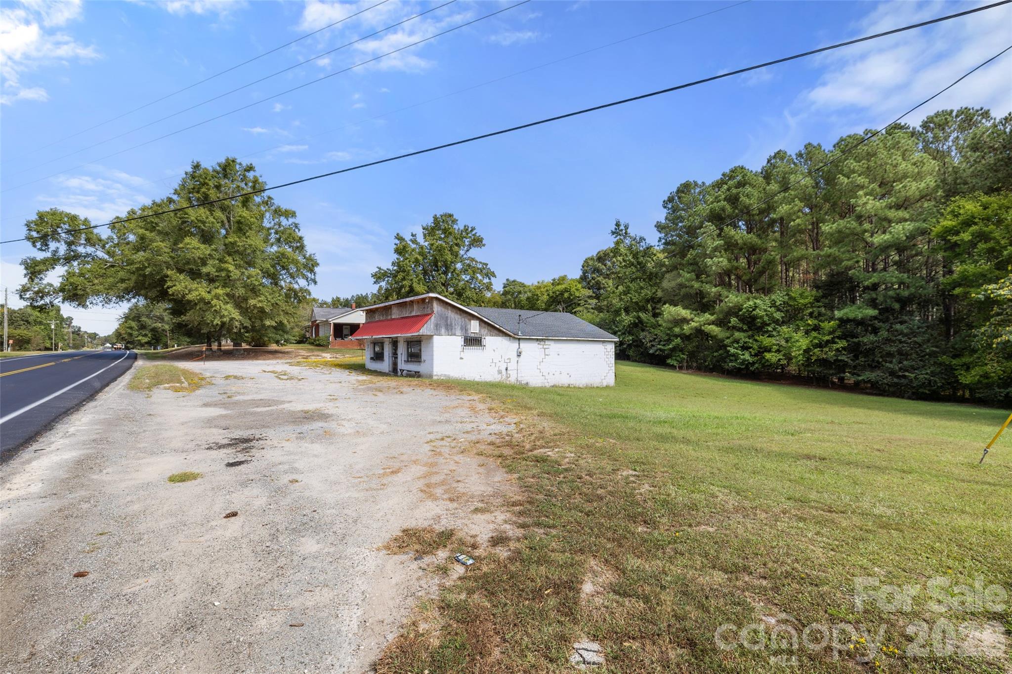 Small white roadside building with a red awning, gravel parking area, and wooded lawn.